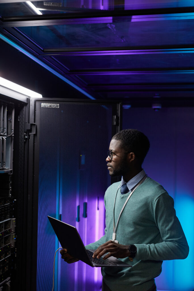 When to add programmatic - Vertical portrait of young African American data engineer holding laptop while working with supercomputer in server room lit by blue light, copy space